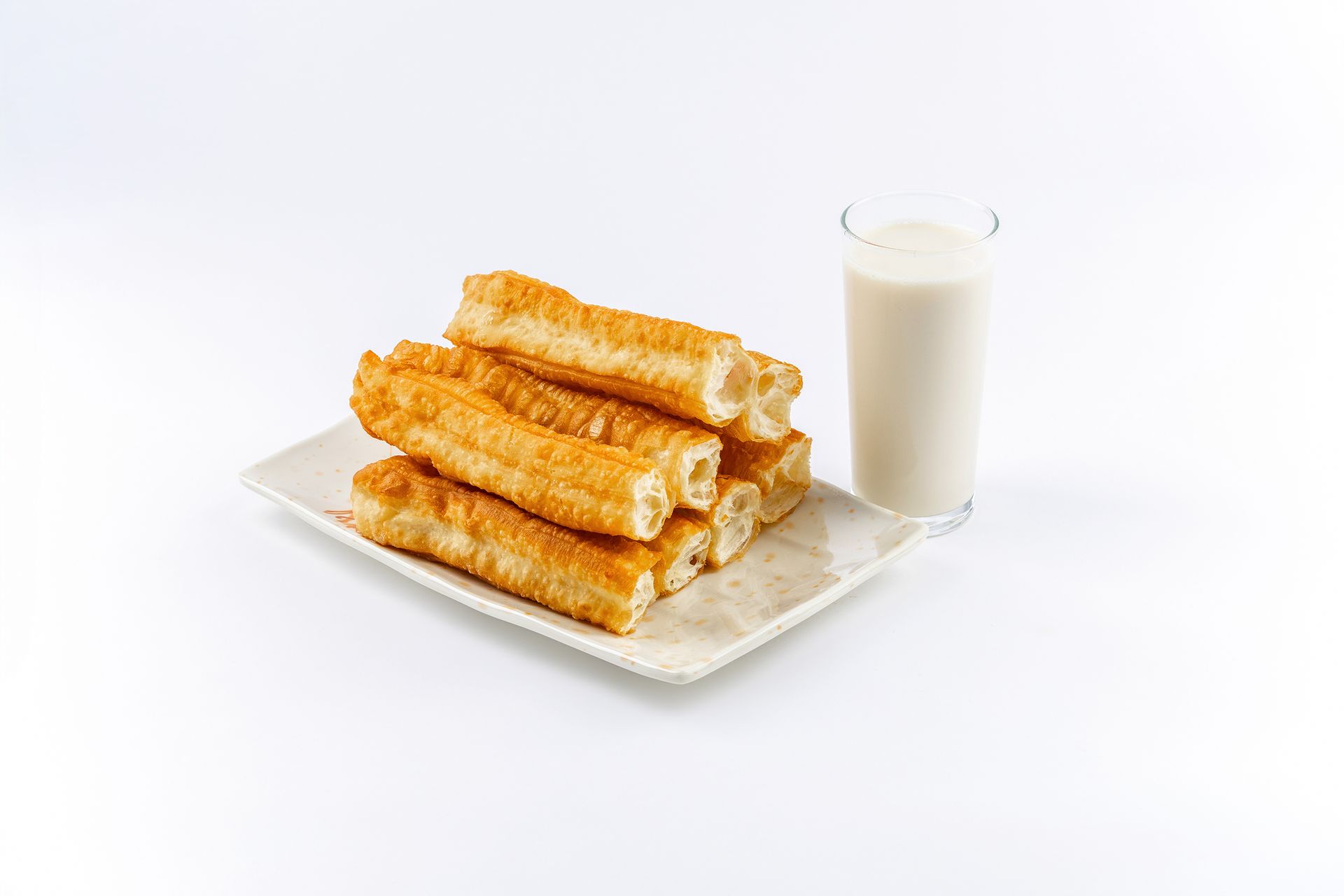 A plate of golden-brown fried dough sticks alongside a glass of soy milk on a white background.