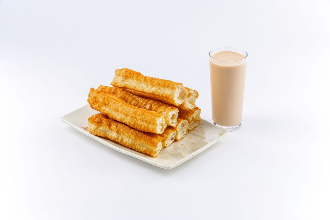 A plate of golden-brown Chinese fried dough sticks, known as youtiao, served next to a glass of soy milk.