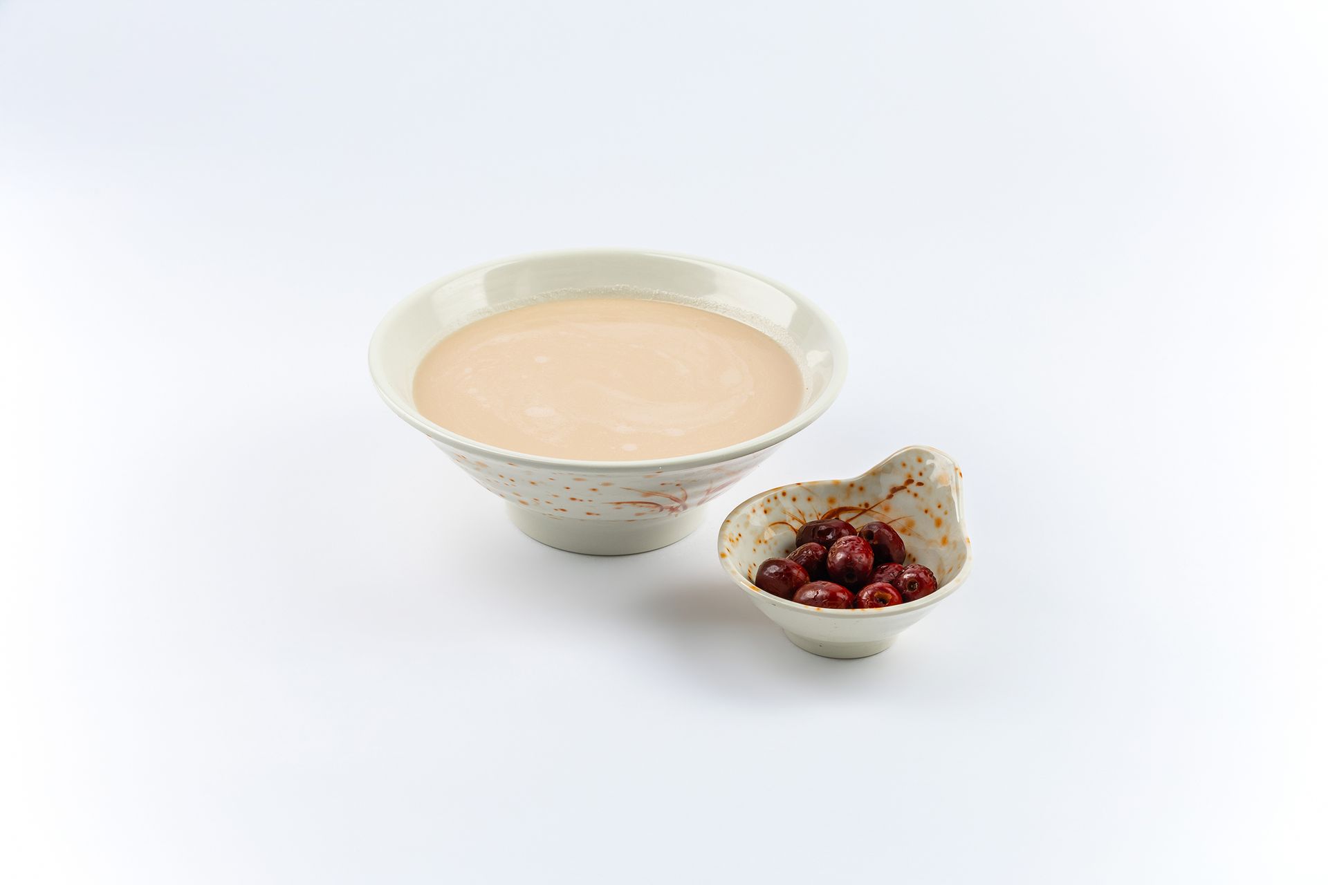 A bowl of pale peach-colored soup next to a small dish of red cherries on a white background.