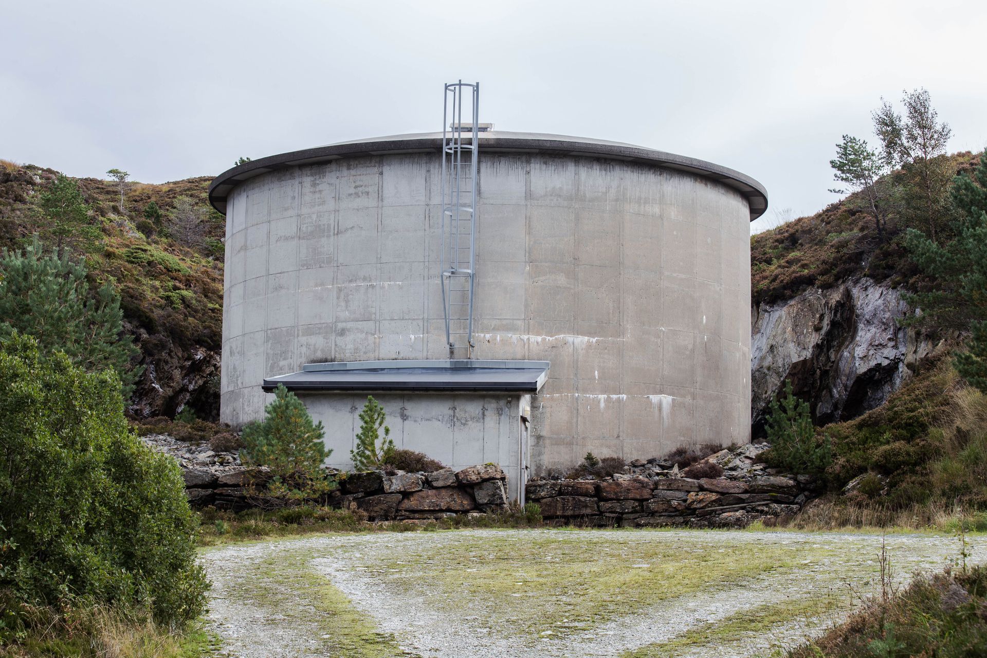 Large concrete water tank surrounded by lush green trees and hills under a clear blue sky.