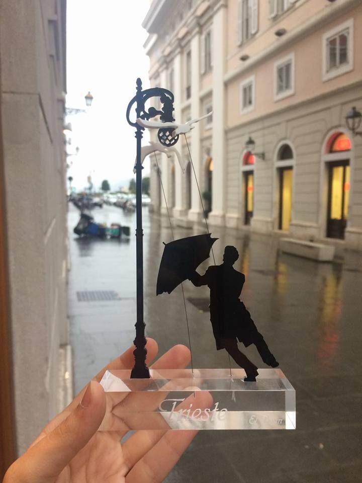 Mano che regge un souvenir di Trieste: la sagoma di una persona con un ombrello, accanto a un lampione, su una strada bagnata.
