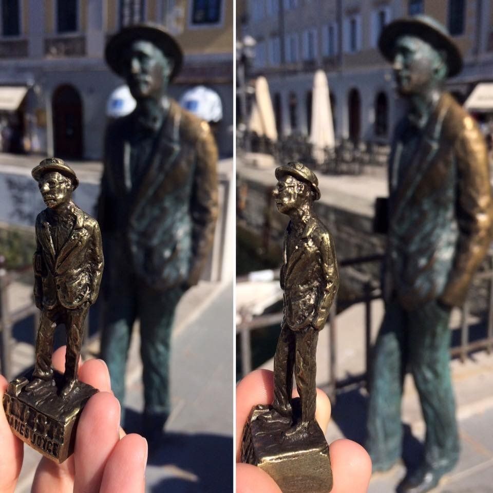 Statua in bronzo raffigurante un uomo che tiene in mano una replica in miniatura di fronte a una statua più grande, all'aperto.