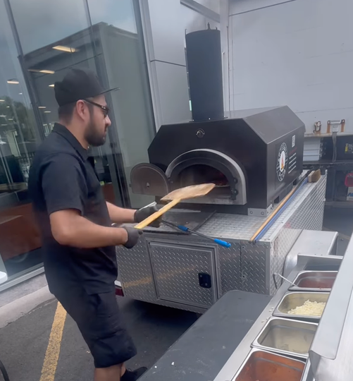 Man making pizza with a mobile oven, in an outdoor setting.