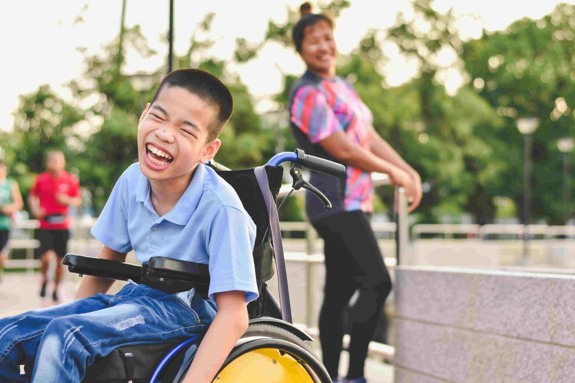 Boy in wheelchair laughing outdoors; woman stands in background.