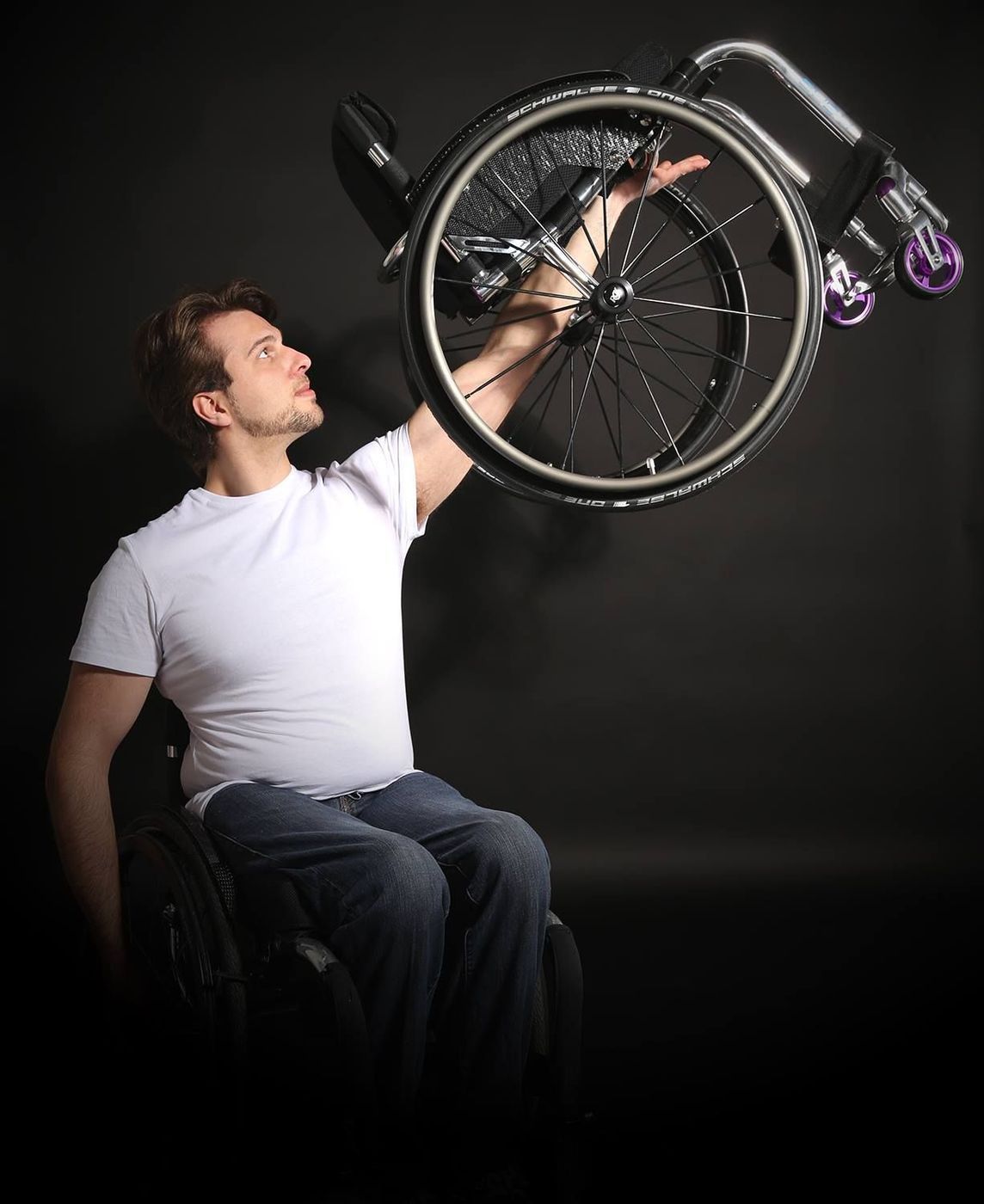 Man in wheelchair holding up a wheelchair wheel, looking up. Black background.