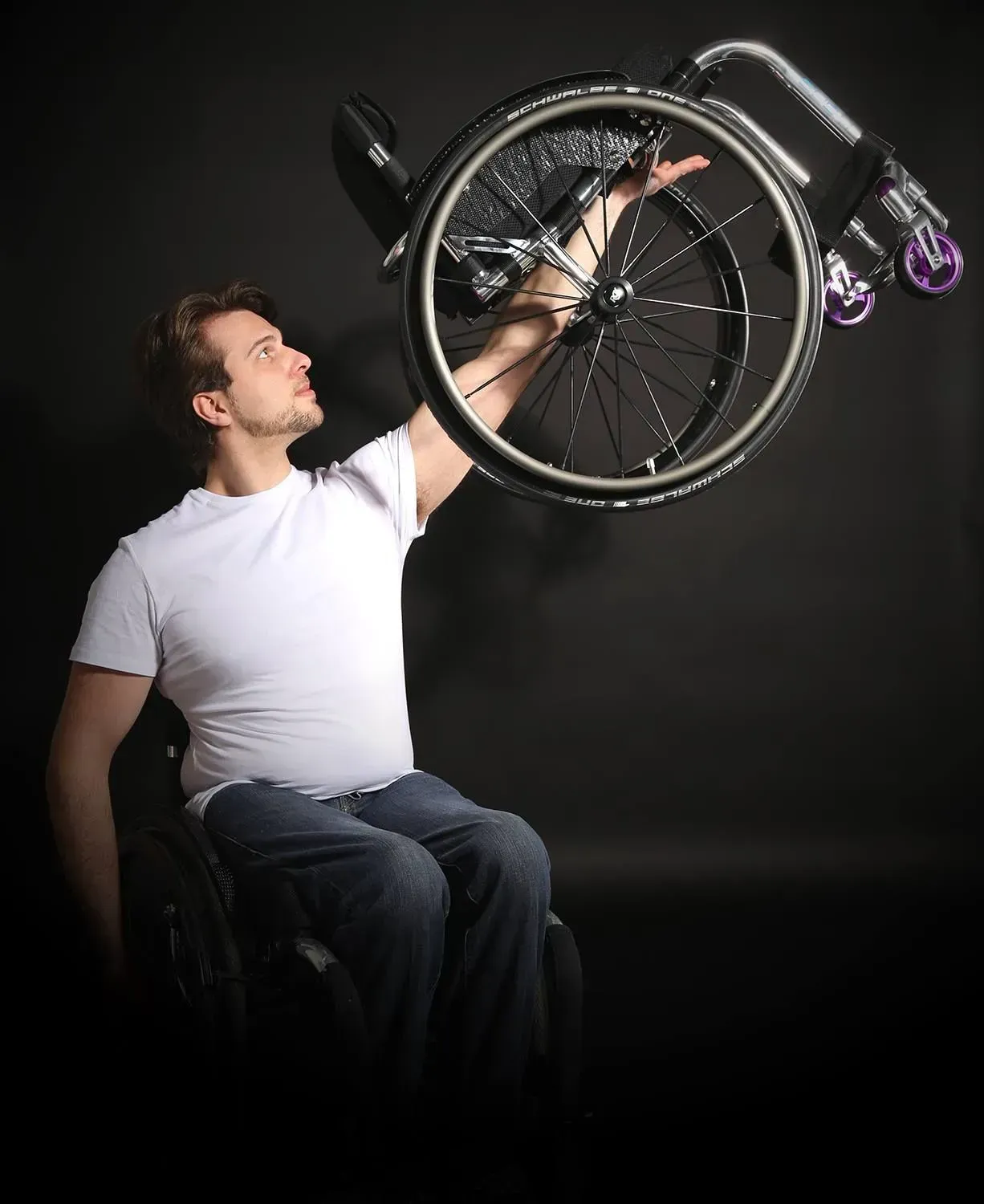 Man in wheelchair holding up a wheel, looking upwards, dark background.