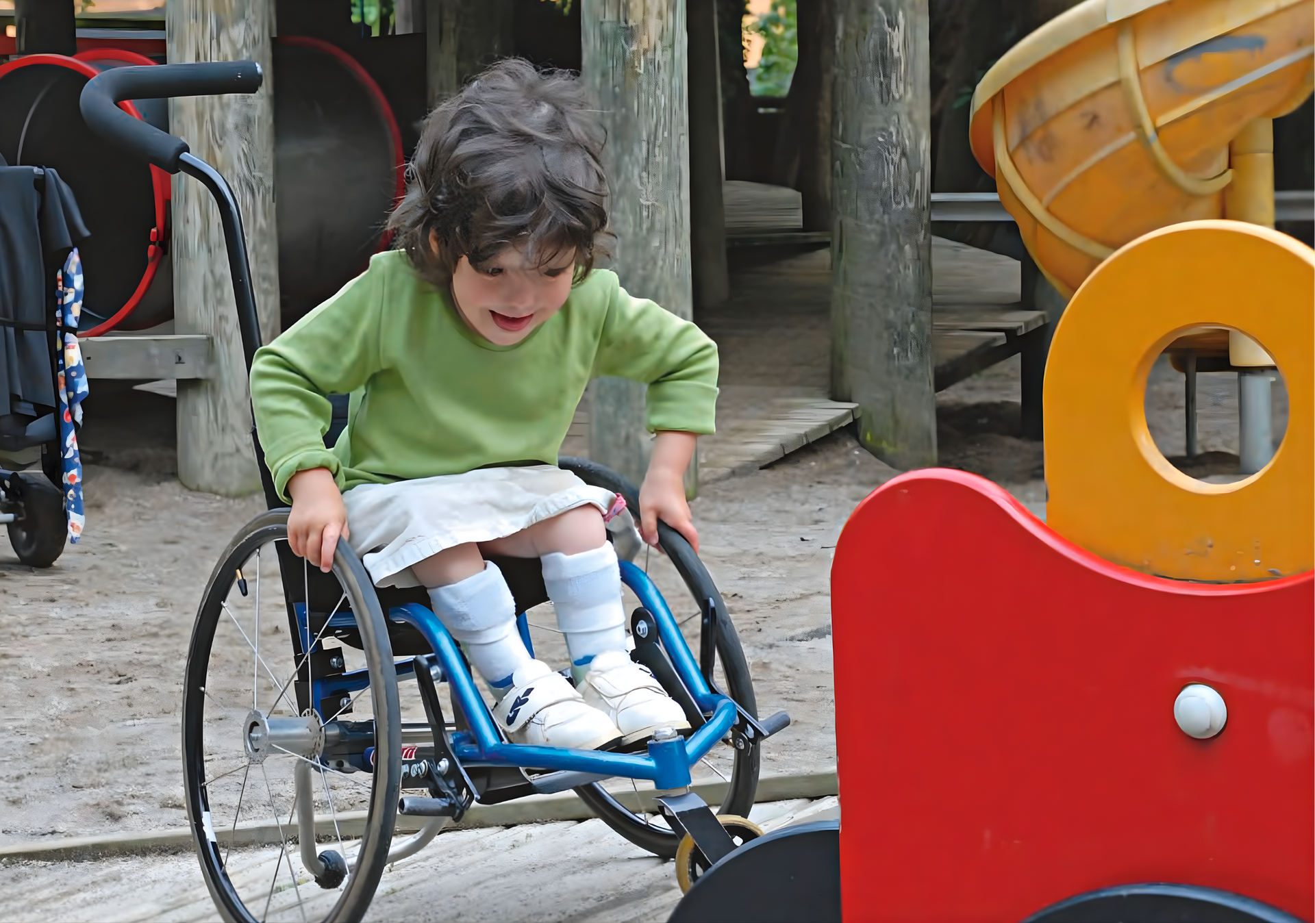 Child in a wheelchair at a playground, wearing a green shirt and white skirt, looking down with a smile.