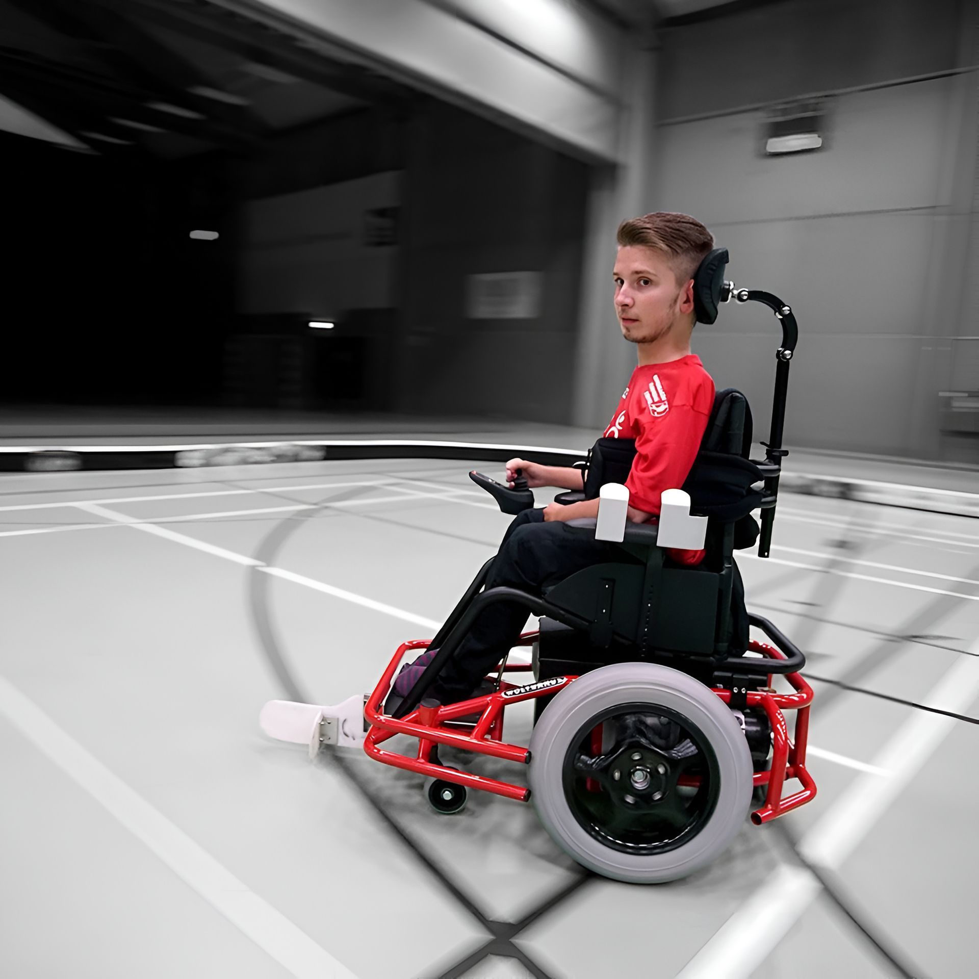 Young man in a red and black wheelchair playing powerchair football on an indoor court.