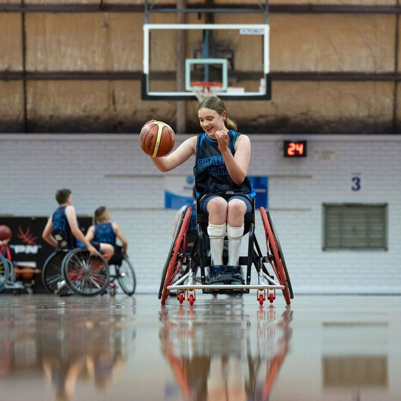 Woman in wheelchair playing basketball, dribbling ball on court, smiling.