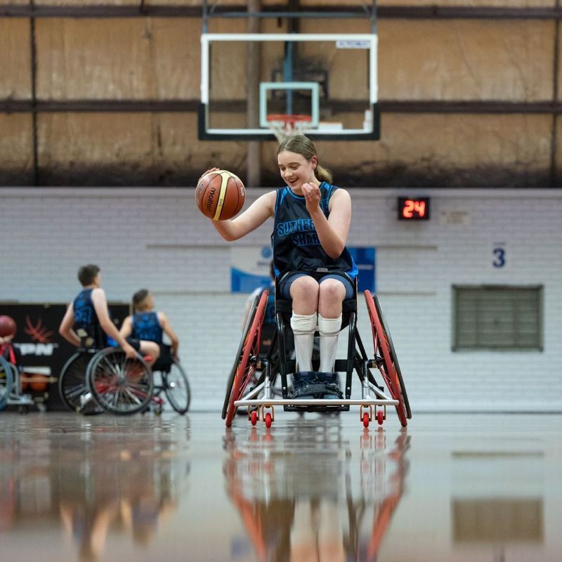 Woman in wheelchair dribbles basketball on court, smiling. Teammates in background.