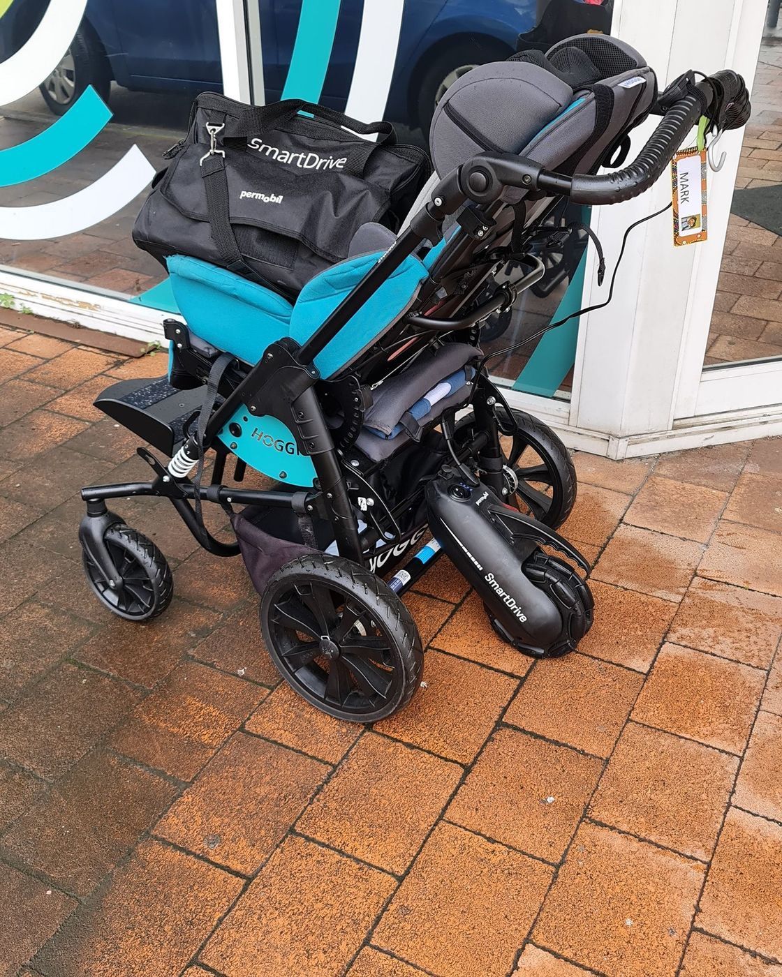 Blue and black adaptive stroller with a black bag on a brick sidewalk.