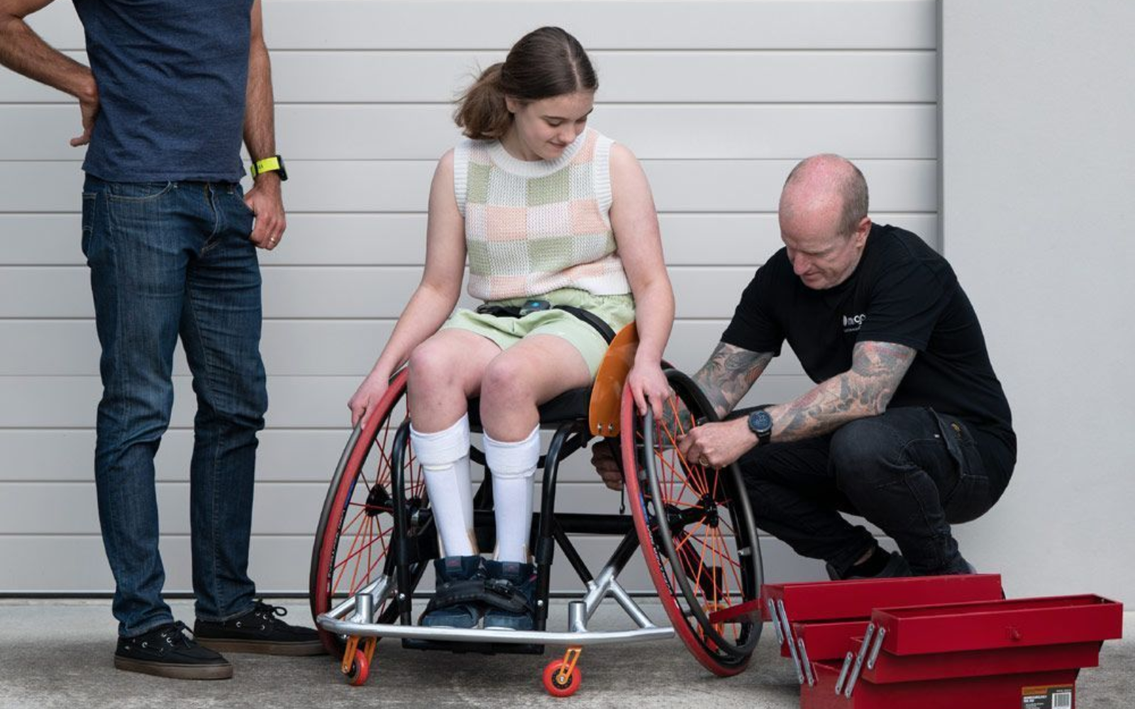 Woman in wheelchair, two men. One man adjusts a wheel. Outdoors, garage door.