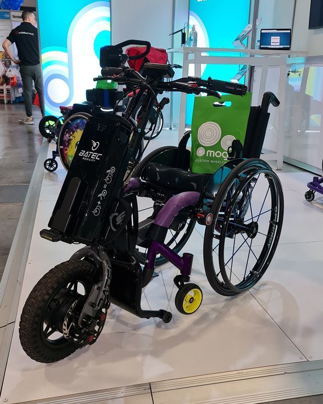 A modified wheelchair with a front wheel motor, purple accents, and black seat; indoor setting.