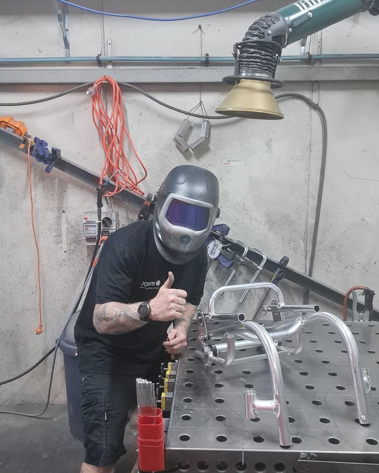 Welder giving thumbs up next to a wheelchair frame, wearing helmet in a workshop.