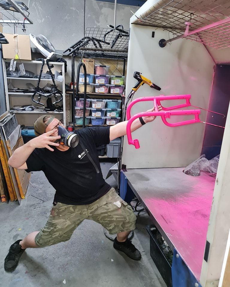 Man in respirator sprays neon pink paint inside booth, holding wheelchair frame.