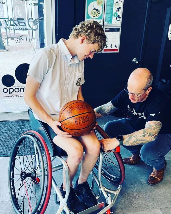 Young man in a wheelchair holds a basketball; man helps him adjust his chair. Indoors, storefront background.