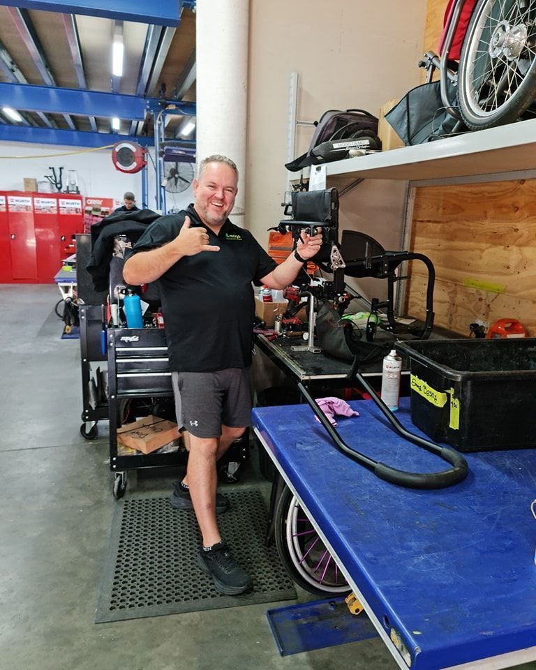 Man in black shirt and shorts gives shaka sign in a workshop with tools and a wheelchair.
