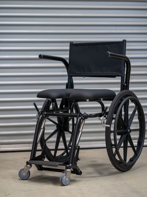 Black wheelchair with black seat and back, against a grey corrugated metal background.