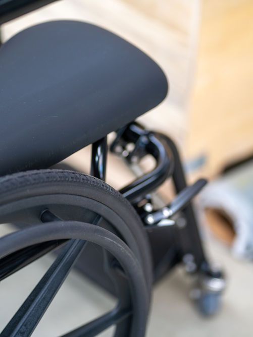 Close-up of a black wheelchair: seat and wheel visible, black frame, blurry background.