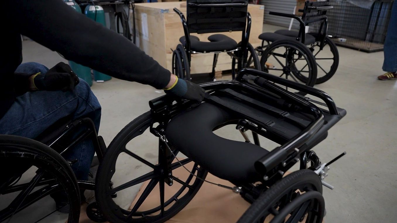 Person folding a black wheelchair indoors, other wheelchairs in background.
