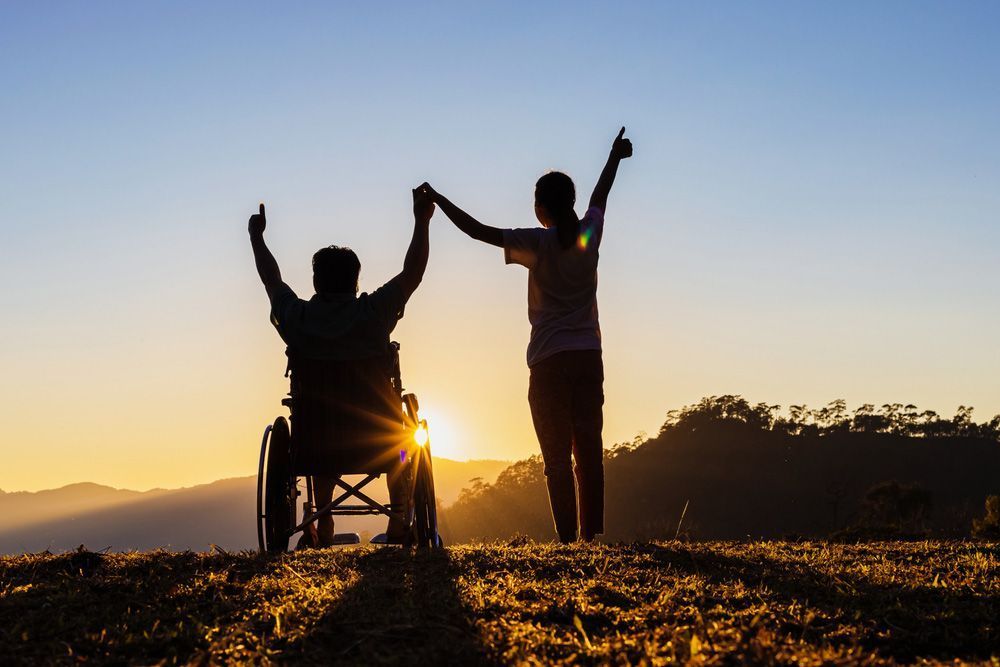Silhouette of person in wheelchair and another person holding hands, arms raised, against a sunset backdrop.