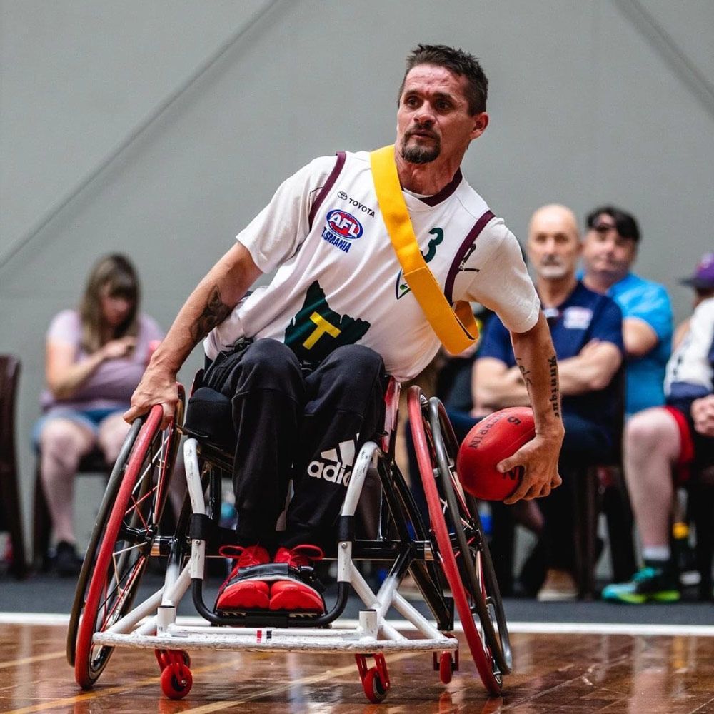 Man in wheelchair playing wheelchair Australian rules football, holding a red ball, in a gym.