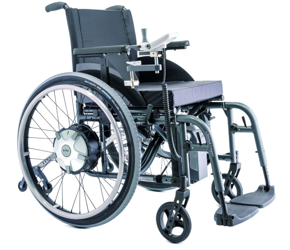 Electric wheelchair, gray frame, black seat and armrests, control arm, large rear wheels, small front wheels, indoor setting.