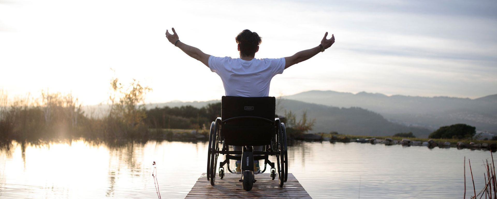 Person in a wheelchair with arms raised, on a wooden dock overlooking a lake and mountains.