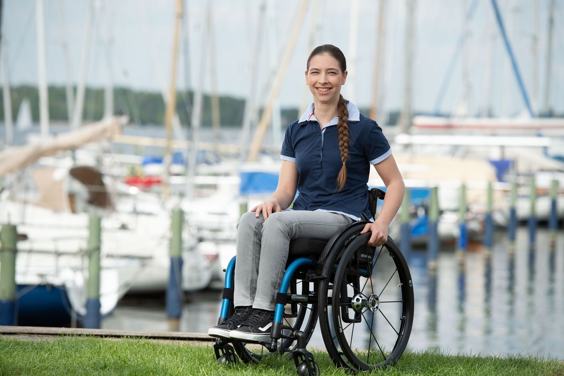 Woman in wheelchair smiling at a marina, wearing blue polo shirt and gray pants.