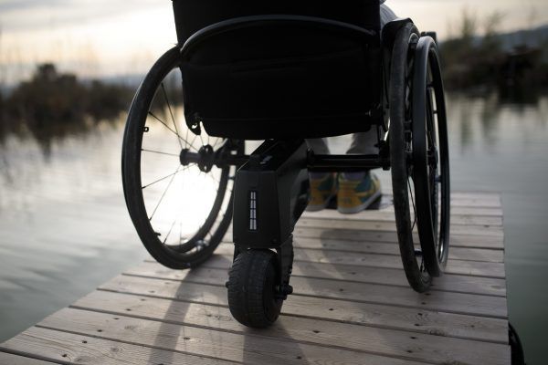 Wheelchair on a wooden dock overlooking water. Black chair, focus on wheels. Setting is outdoors.