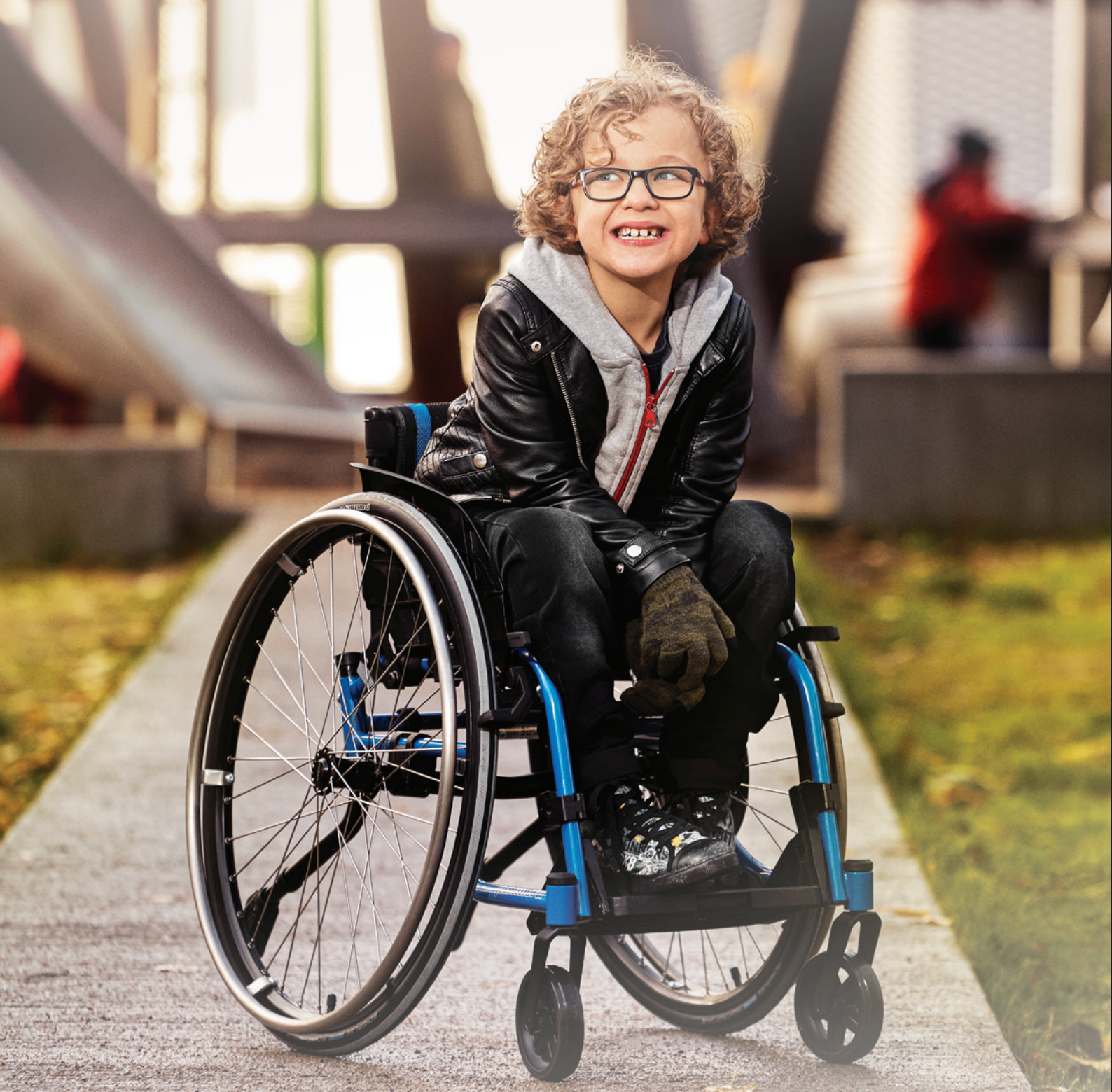 Boy in wheelchair smiles outdoors, wearing glasses and jacket. Pathway, park setting.