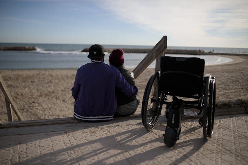 Two people sit on a beach, gazing at the sea. A wheelchair sits nearby on a walkway. Sunny day.