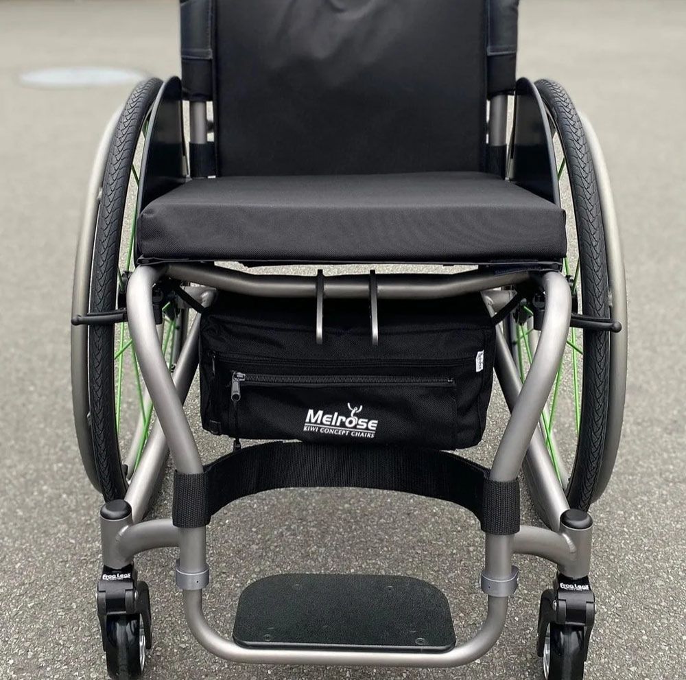 A close-up of a gray wheelchair on pavement; features include black seat, wheels with green accents, and a black storage bag.