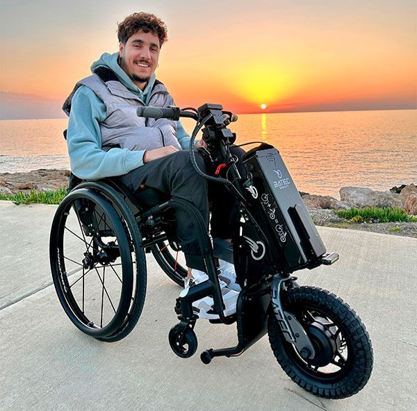 Man in wheelchair with attached front-end, smiles at sunset over ocean.