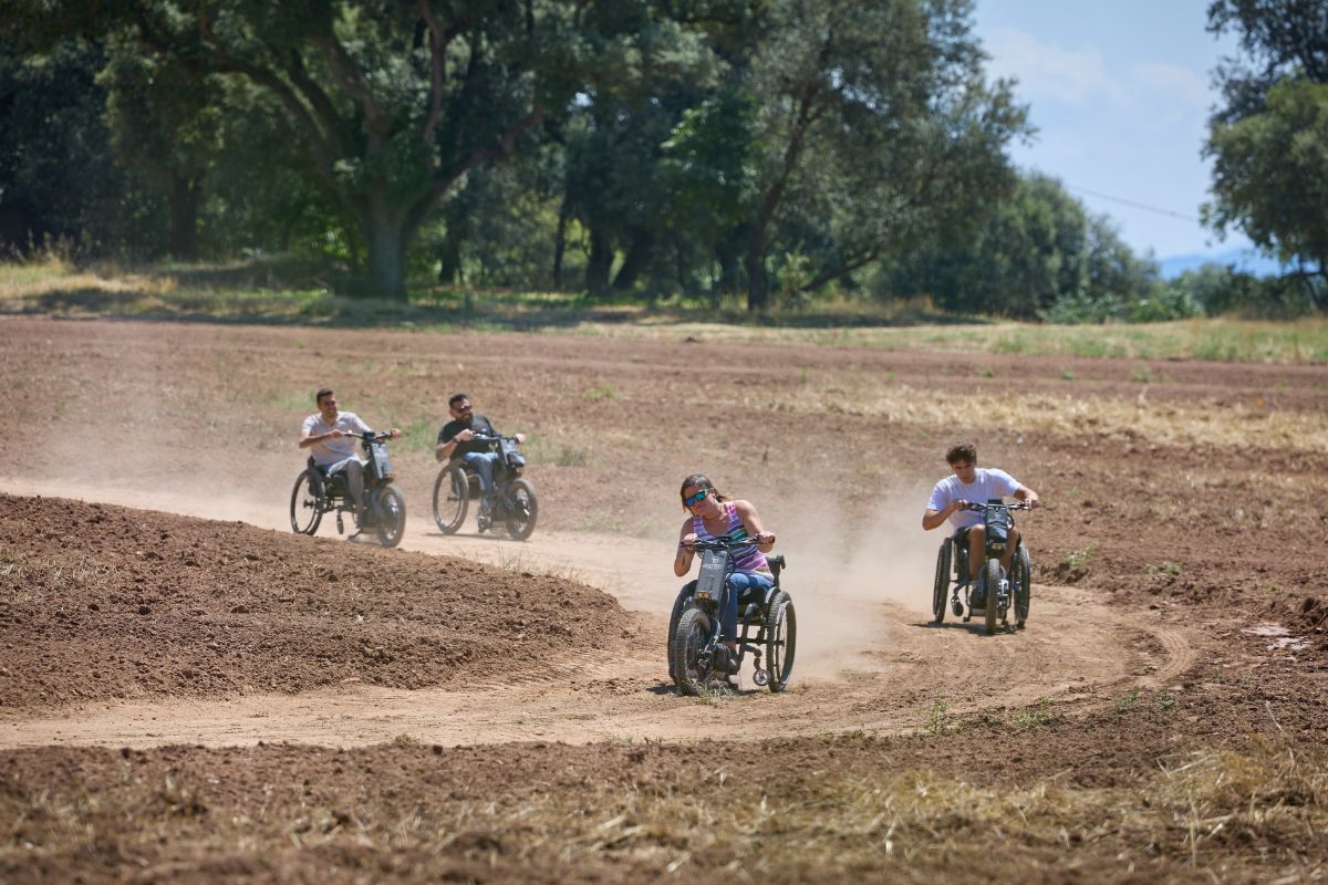 Four people in wheelchairs racing on a dirt track, kicking up dust under a sunny sky.