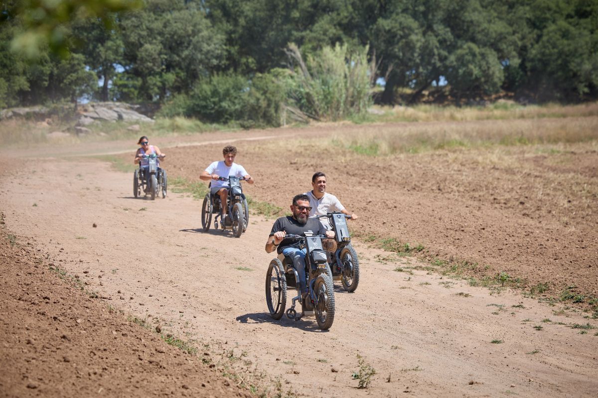 Four people ride motorcycles on a dirt path in a sunny, rural area, kicking up dust.