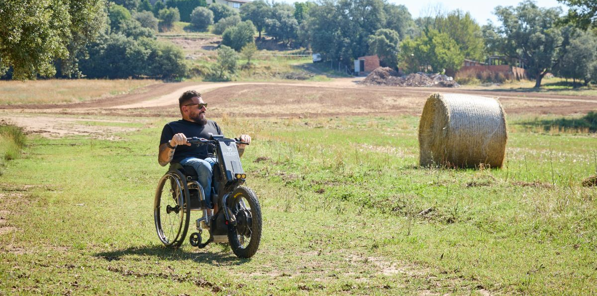 Man in wheelchair on a grassy field near a hay bale, trees, and buildings on the horizon.