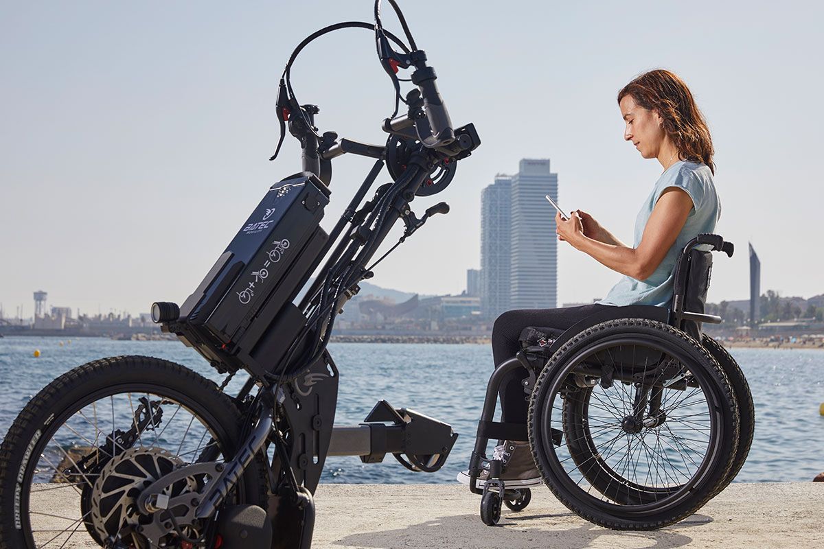 Woman in wheelchair with electric handcycle by water, using a phone.