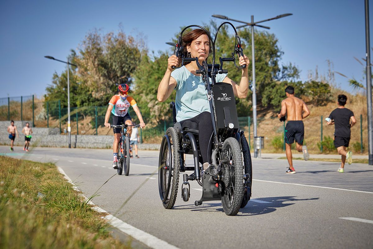 Woman in a wheelchair smiles, using a hand-cycle on a paved path, with cyclists and runners in the background.