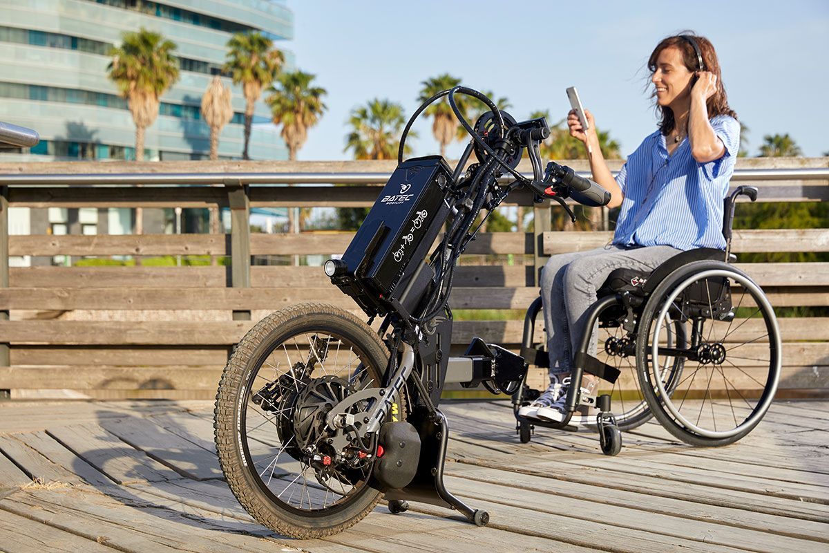 Woman in wheelchair on boardwalk using attached electric bike, looking at phone, sunny day.