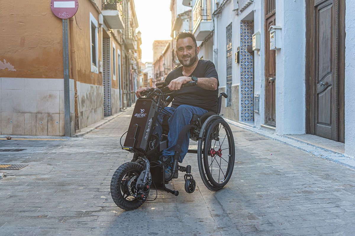 Man in a wheelchair smiles on a cobblestone street, sunlit. He sits with his hands on the wheelchair handles.