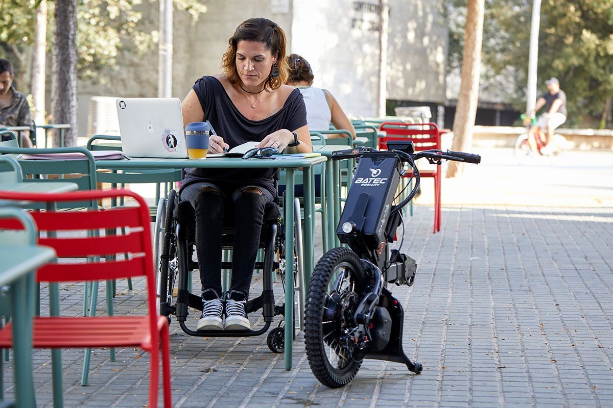 Woman in wheelchair working at an outdoor table with a laptop, next to a mobility device.