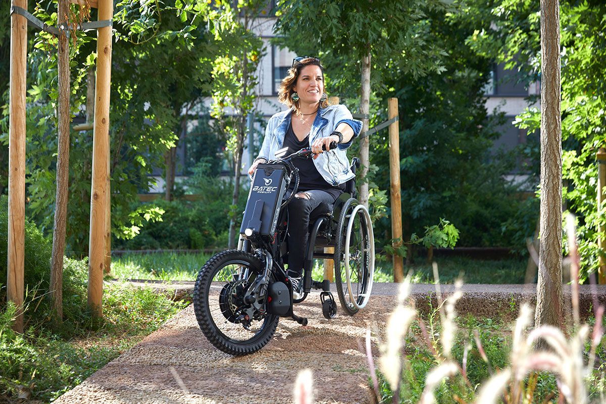 Woman in a wheelchair, smiling, navigating a stone path in a park, trees surround her.