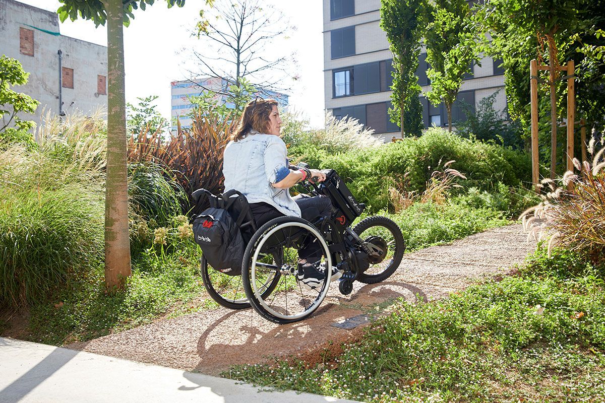 Woman using a three-wheeled wheelchair on a gravel path surrounded by greenery.