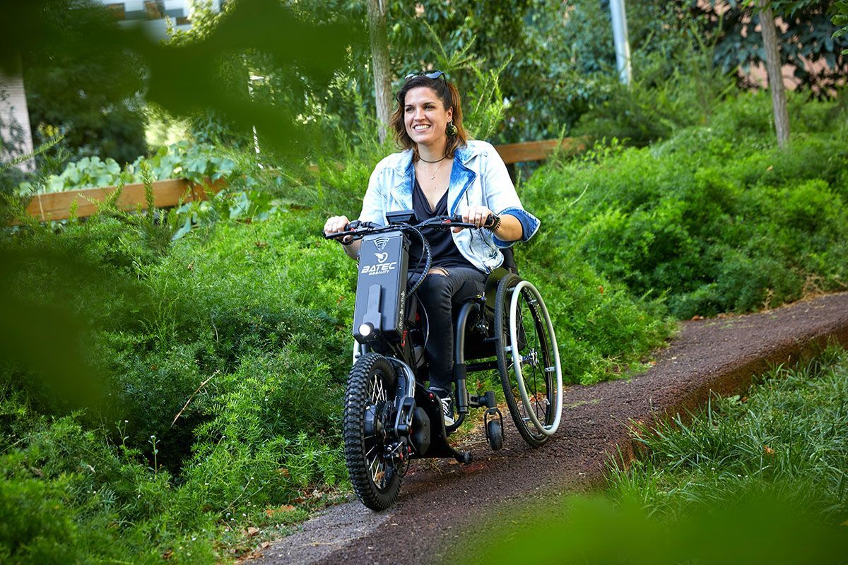 Woman using a motorized wheelchair on a wooded path, smiling.