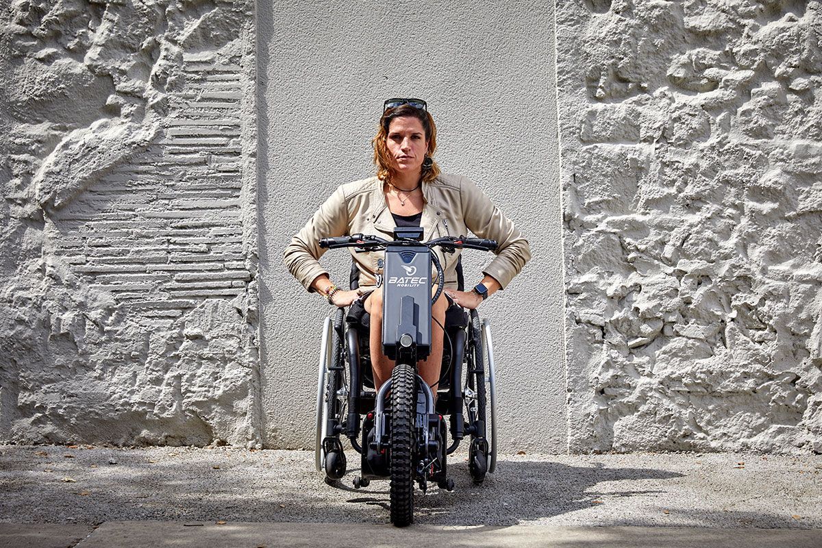 Woman in wheelchair with an electric handcycle in front of a textured stone wall.