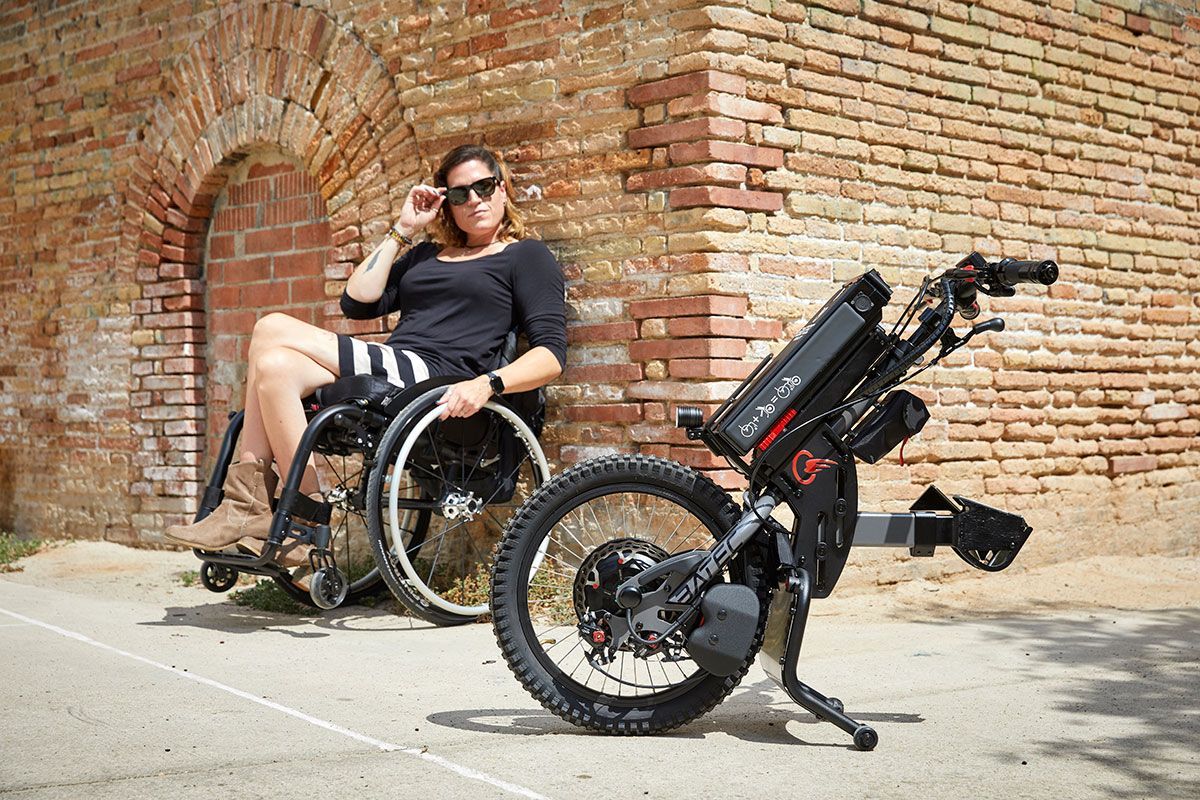 Woman in wheelchair with electric handcycle outdoors near brick wall.