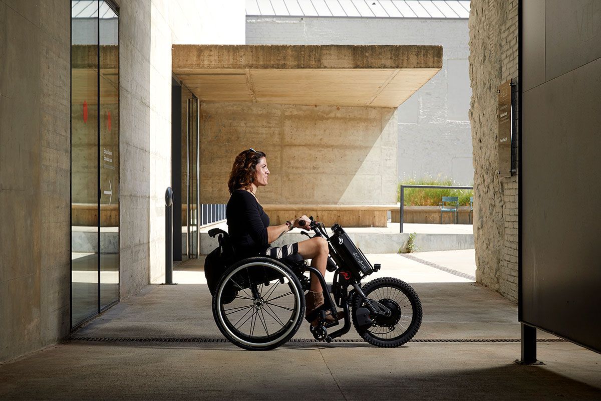 Woman in wheelchair outside a building. Concrete architecture and bright sunlight.
