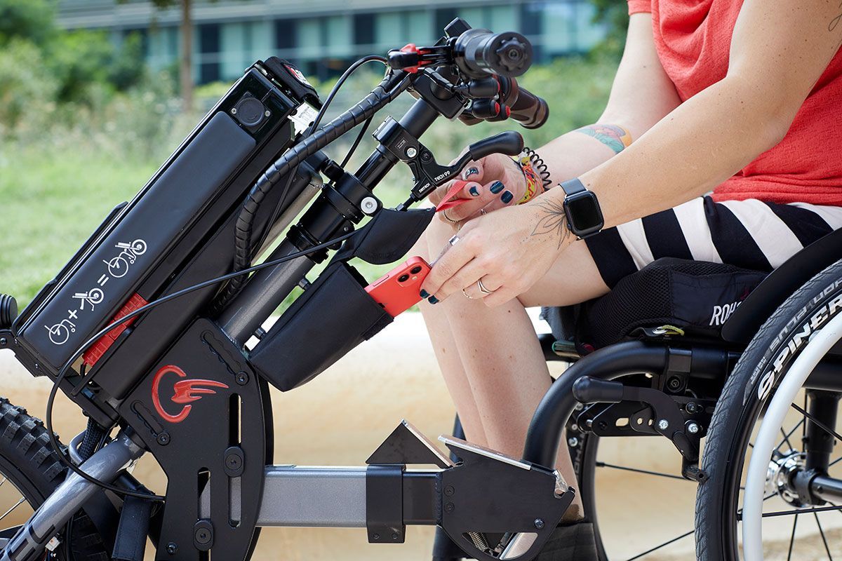 Person in a wheelchair using controls on a black and red motorized attachment outdoors.