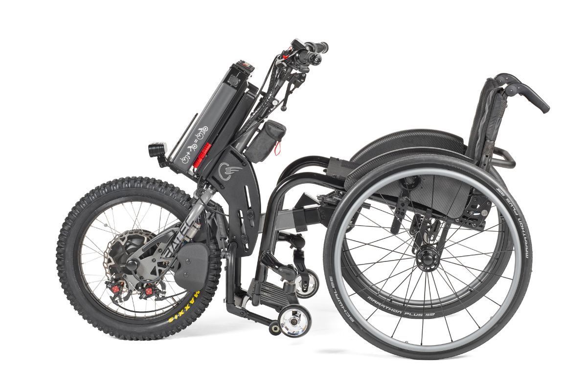 Motorized wheelchair with large front tire, side view. Black frame and components, battery pack visible.
